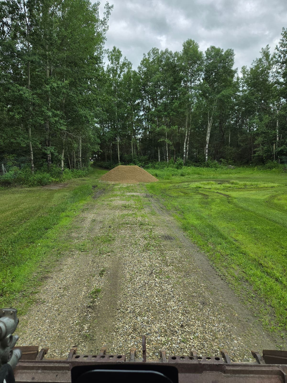 View from the cab of the track loader down a grass two-track, with a fresh pile of gravel dropped at the far end
