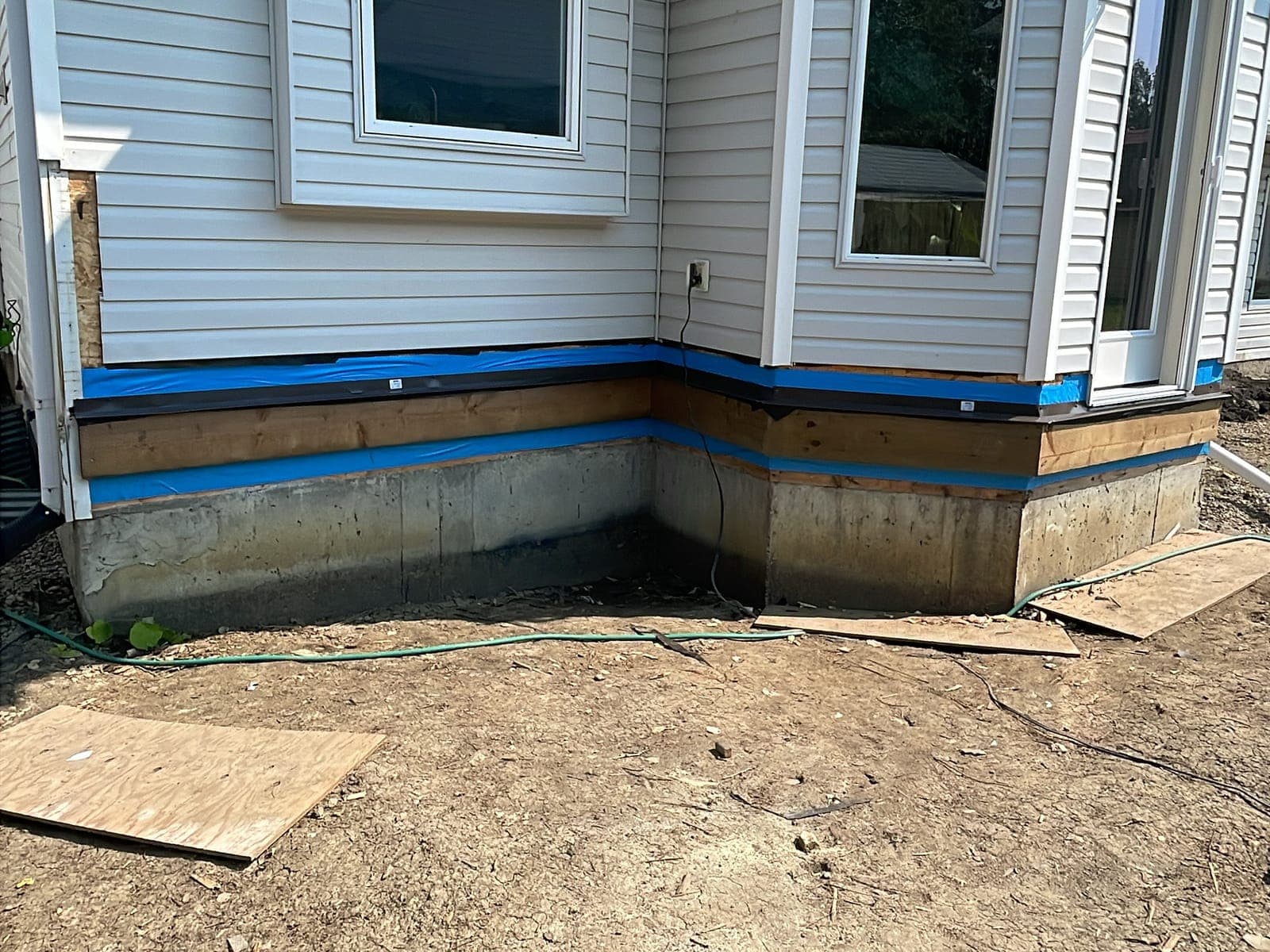 Exposed concrete house foundation with bare soil against it and plywood on the ground